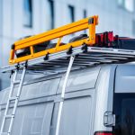 A van with a ladder rack shows a bright yellow ladder securely mounted on the roof, parked near a modern building.